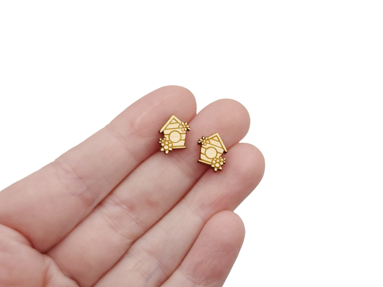 a hand holding a pair of wooden cabochon stud earring blanks cut and engraved to look like a birdhouse with flowers