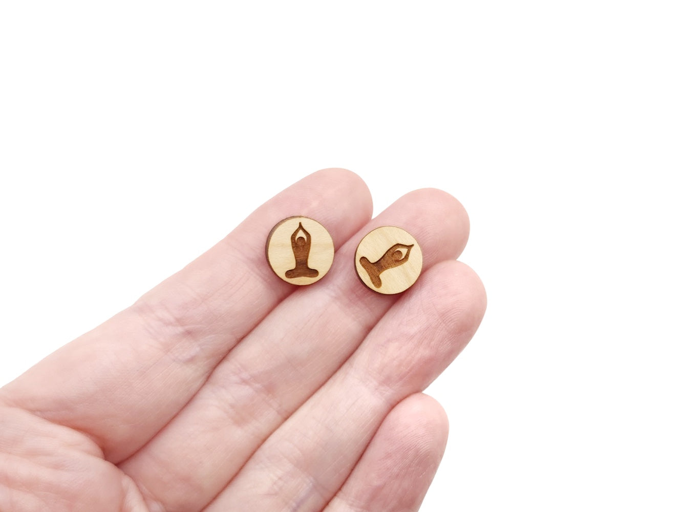 a hand holding a pair of round wooden cabochon earring blanks engraved with a yoga pose silhouette design