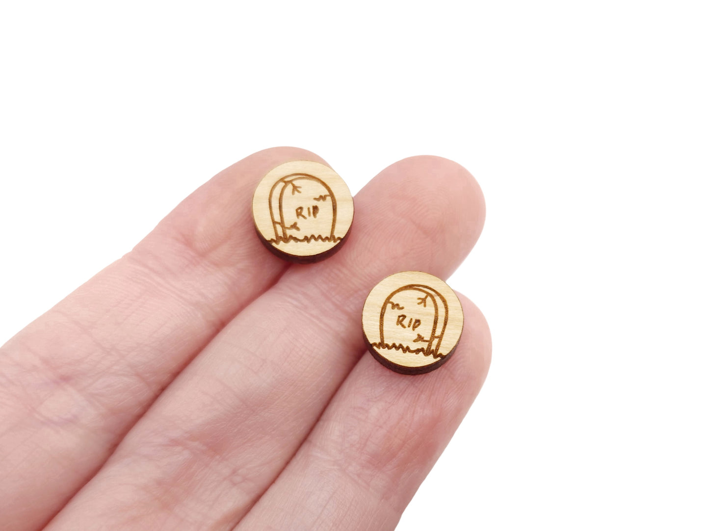 a hand holding a pair of round wooden cabochon earring blanks engraved with a Halloween Tombstone