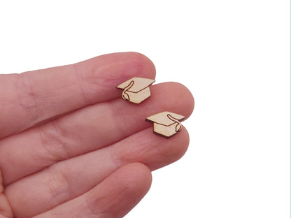 hand holding a pair of small wooden cabochon blanks cut and engraved to look like graduation caps