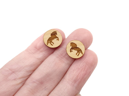 a hand holding a pair of round wooden cabochon earring blanks engraved with a rearing horse silhouette design