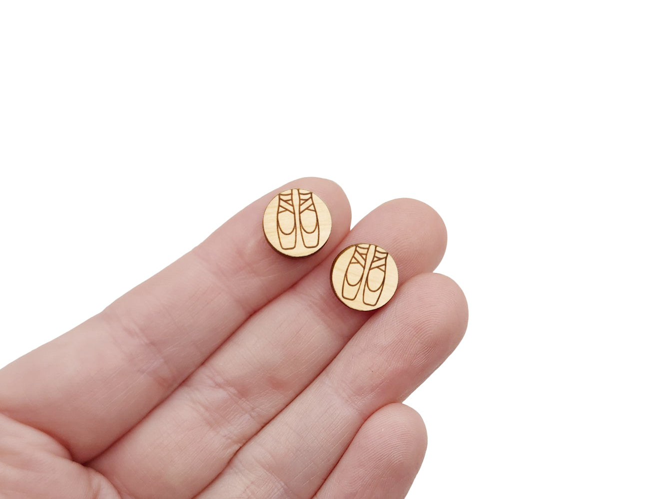 a hand holding a pair of round wooden cabochon earring blanks engraved with a pair of ballet slippers