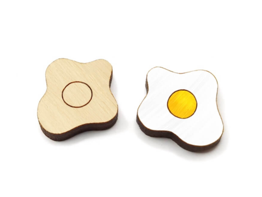 a pair of wooden cabochon stud earring blanks cut and engraved to look like fried eggs, one is painted