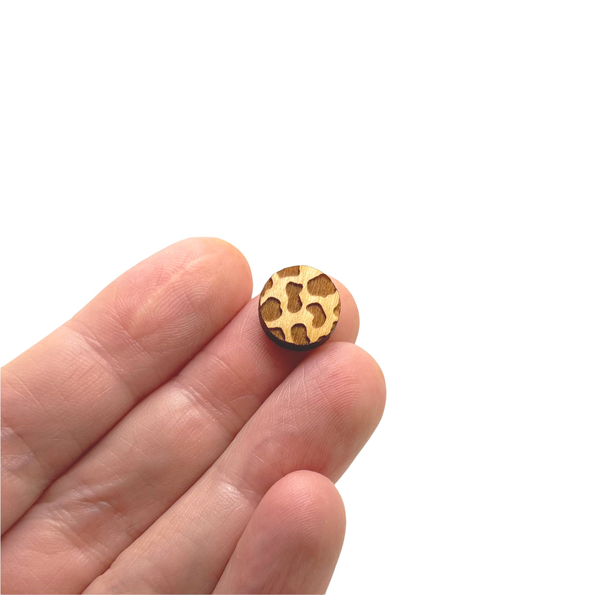 a hand holding a pair of round shaped wooden cabochon stud earring blanks engraved with cow print